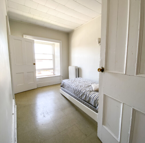 An open door reveals a bedroom with a window and radiator in this 213 Queen Street unit in downtown Kingston. Bedroom with striped bedding seen through an open door at 213 Queen Street Unit 3, Kingston, Ontario